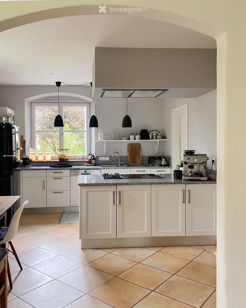 Bright, modern kitchen with white cabinets, granite countertops, and stainless steel appliances. Features include a large window, pendant lights, and a breakfast area with a table and chairs.