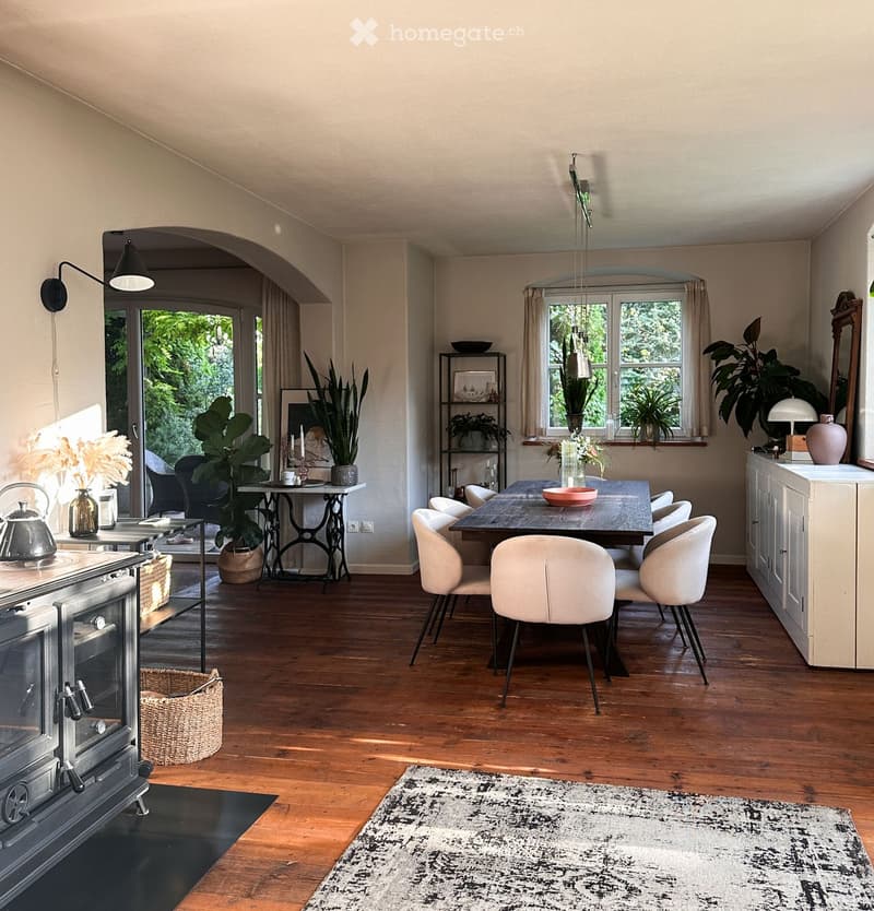 Open-plan kitchen and dining area with wooden flooring, modern furniture, and large windows overlooking greenery.