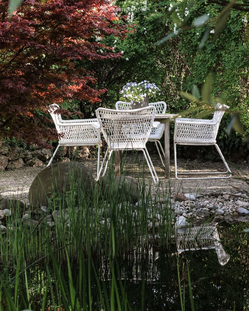 A serene garden area with a small pond, surrounded by lush greenery, featuring a set of white wicker chairs and a matching table.