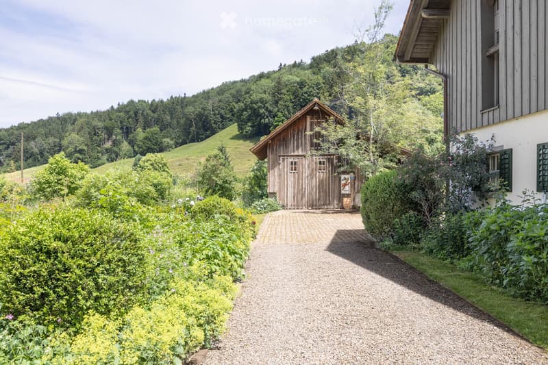 A gravel driveway leads to a wooden barn and a modern house surrounded by lush greenery and flowering bushes.