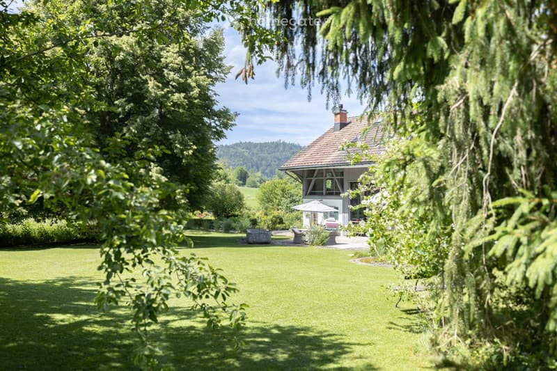 A well-maintained garden with a large lawn, a house with a red-tiled roof, and outdoor seating area with a table and chairs under an umbrella.