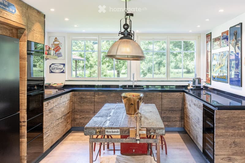 Modern kitchen with wooden cabinets, stainless steel appliances, a large island table, and a hanging pendant light. The space features large windows with a view of greenery outside.