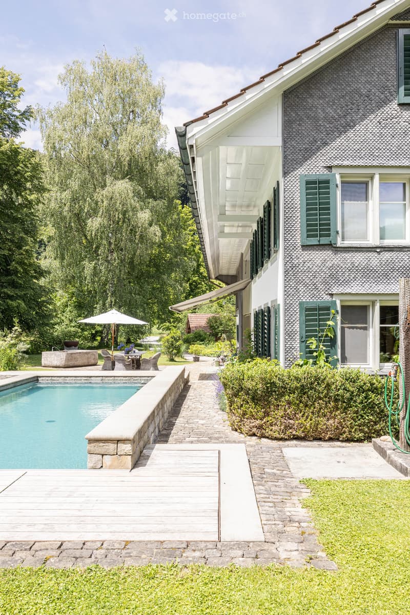 Two-story house with grey shingled roof, white trim, green shutters, and a covered porch. Adjacent to a swimming pool with a stone deck, outdoor seating area, and lush greenery.