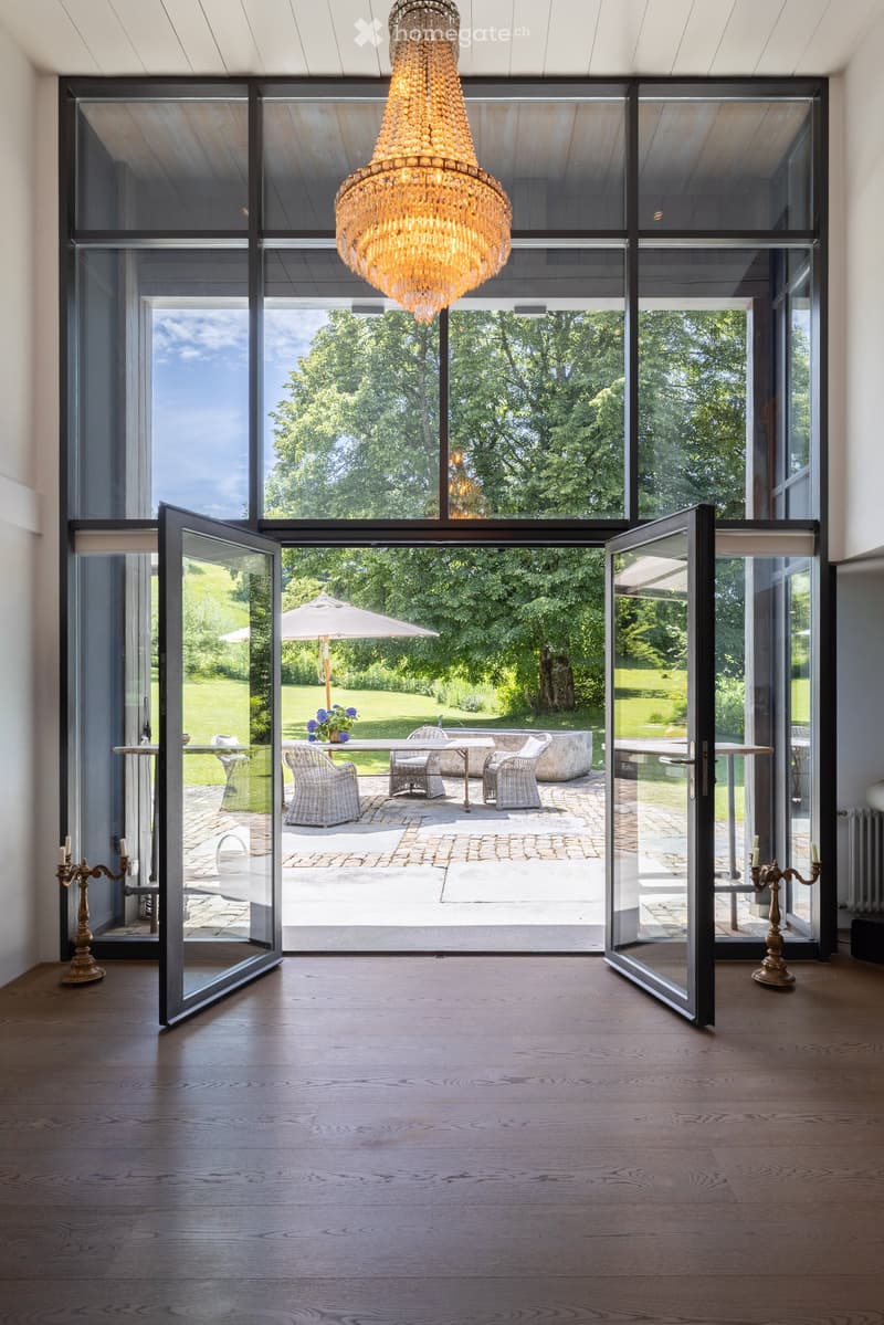 Bright hallway with large glass doors leading to a garden, wooden floor, chandelier, and candle holders on either side.