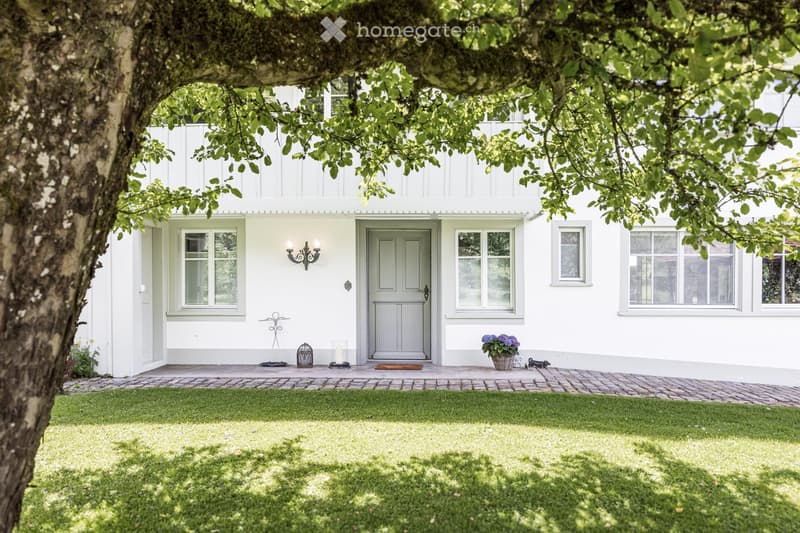Two-story white house with multiple windows, a front door, and a well-maintained lawn. The entrance features a small porch with a doormat, a decorative lantern, and a small garden with a potted plant and a small dog.