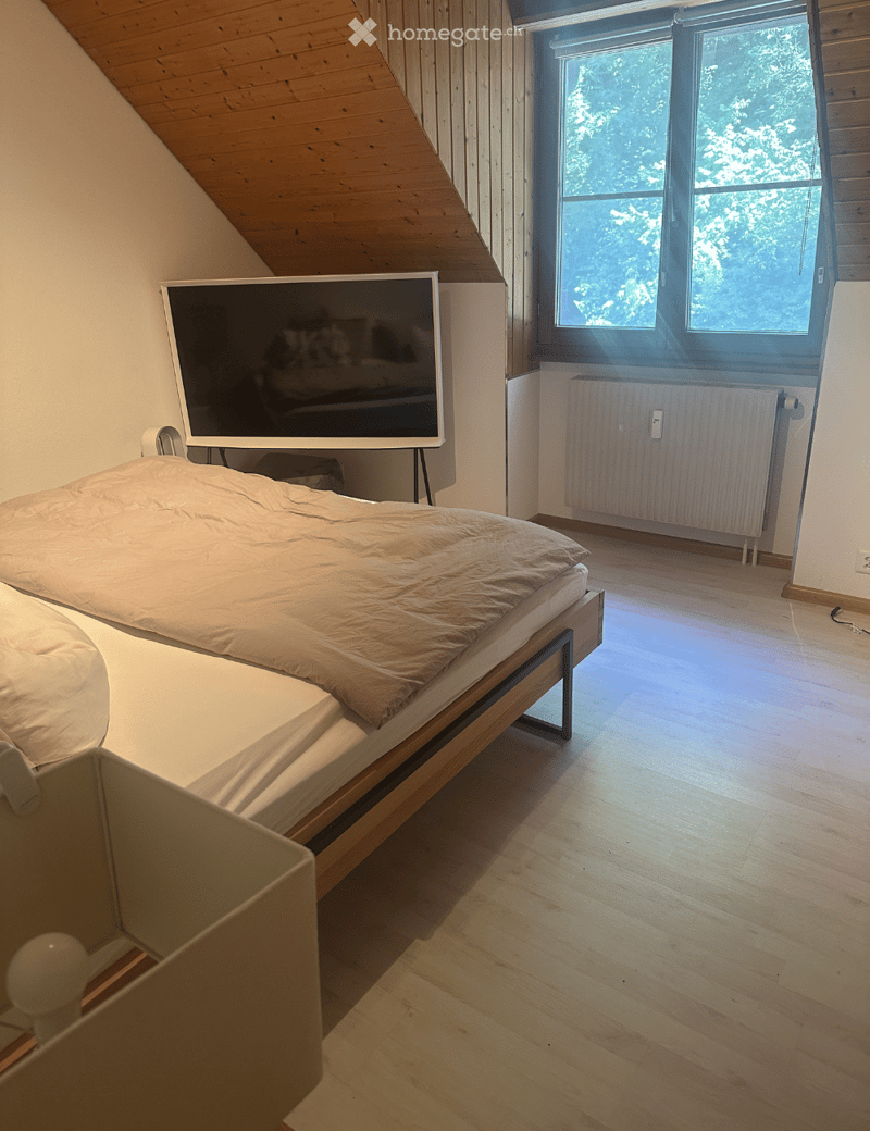 Cozy bedroom with a wooden bed, a television mounted on the wall, and a large window offering a view of greenery.