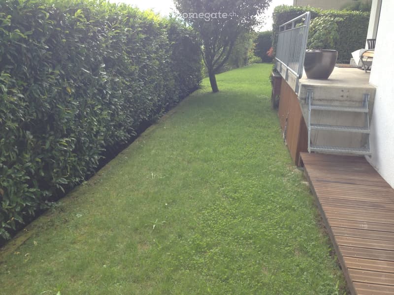 A well-maintained garden with a wooden deck, metal staircase, and a large hedge along the left side.