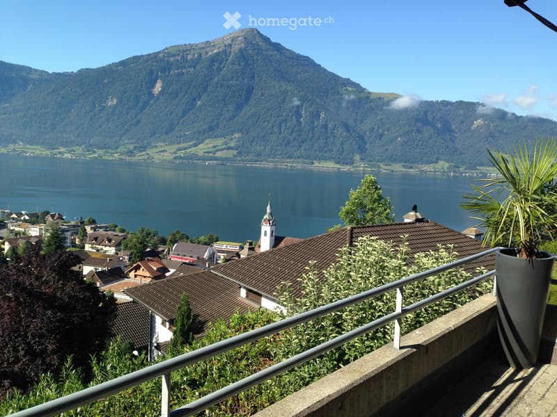 Balcony overlooking a scenic lake and mountain landscape, with a metal railing, potted plants, and a town with a church tower visible in the distance.