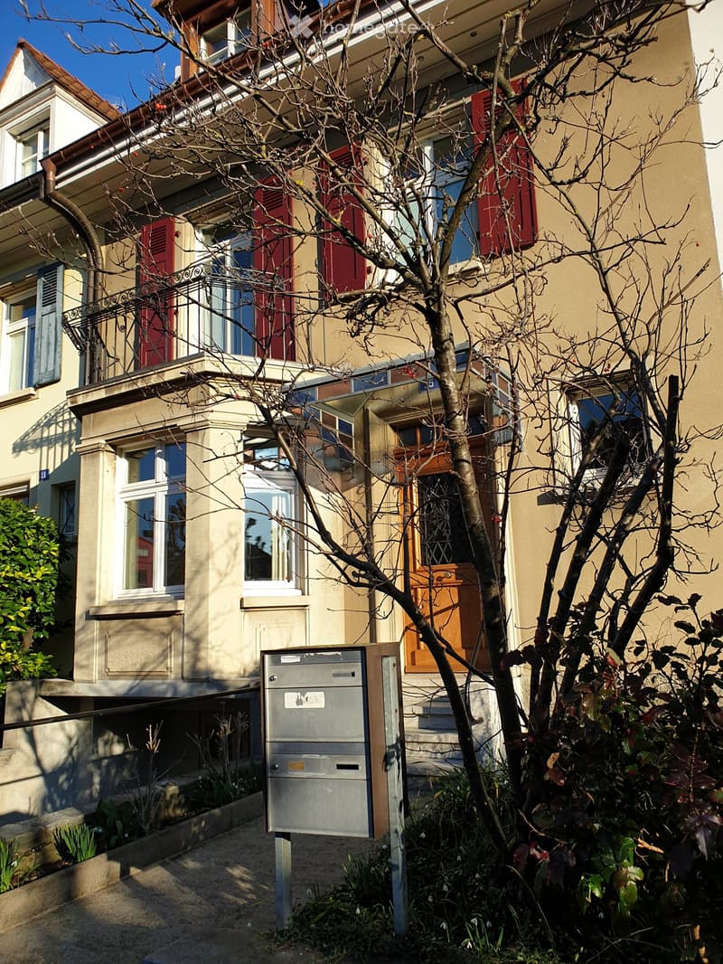 Multi-story residential building with beige and cream exterior, red shutters, and a small garden area with a mailbox and bare tree.