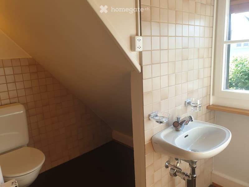 A compact bathroom with a toilet, a wall-mounted sink, and a window providing natural light. The walls are tiled in light beige, and the floor is dark.