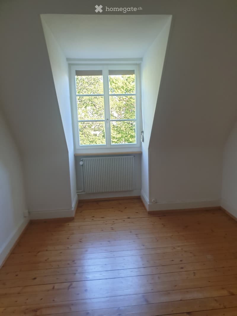 Empty attic room with wooden flooring, white walls, a window with a view of greenery, and a radiator below the window.