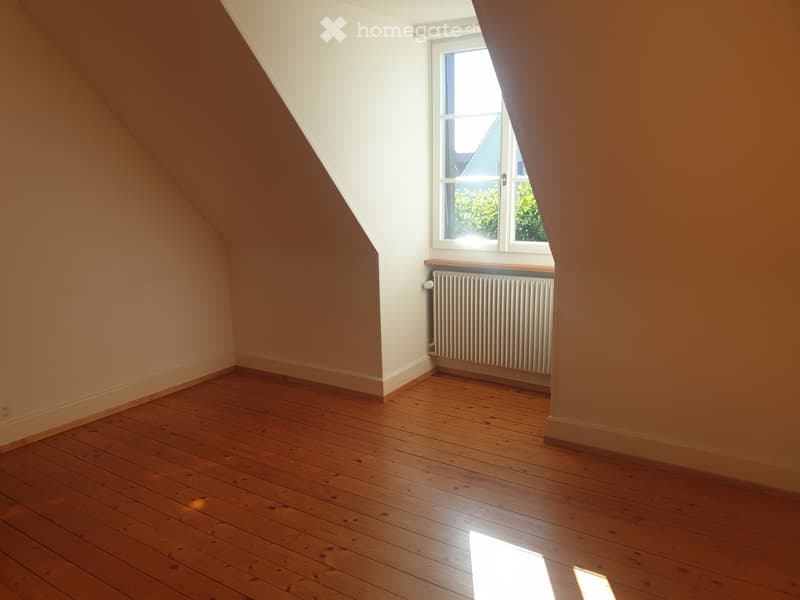 Empty attic room with wooden flooring, white walls, and a single window with a radiator below it.