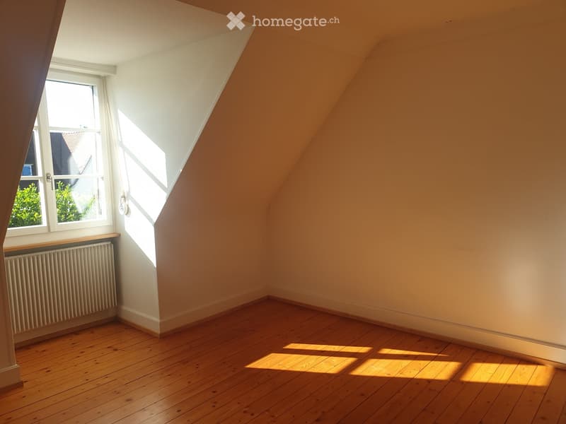 Empty attic room with wooden flooring, slanted ceilings, and a window letting in natural light. The walls are painted in warm tones, and there is a radiator below the window.