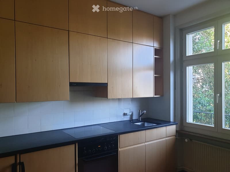 Modern kitchen with light wooden cabinets, black countertop, built-in oven, and a sink with a faucet. Large windows provide natural light.