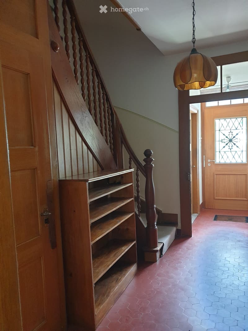 Wooden staircase with turned balusters, wooden shelf, stained glass door, and hanging lamp in a hallway with red and blue tiled floors.