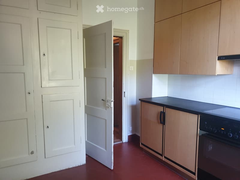 Empty kitchen with wooden cabinets, a black countertop, and a stove. The floor is red tiled, and there are white doors leading to other rooms.