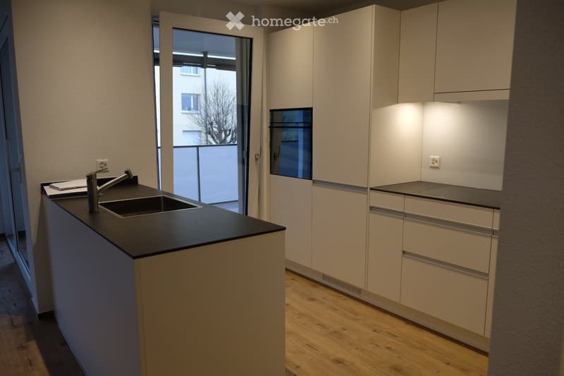 Modern kitchen with light-colored cabinets, black countertop, and a sink with a faucet. The space features wooden flooring and a sliding glass door leading to a balcony.