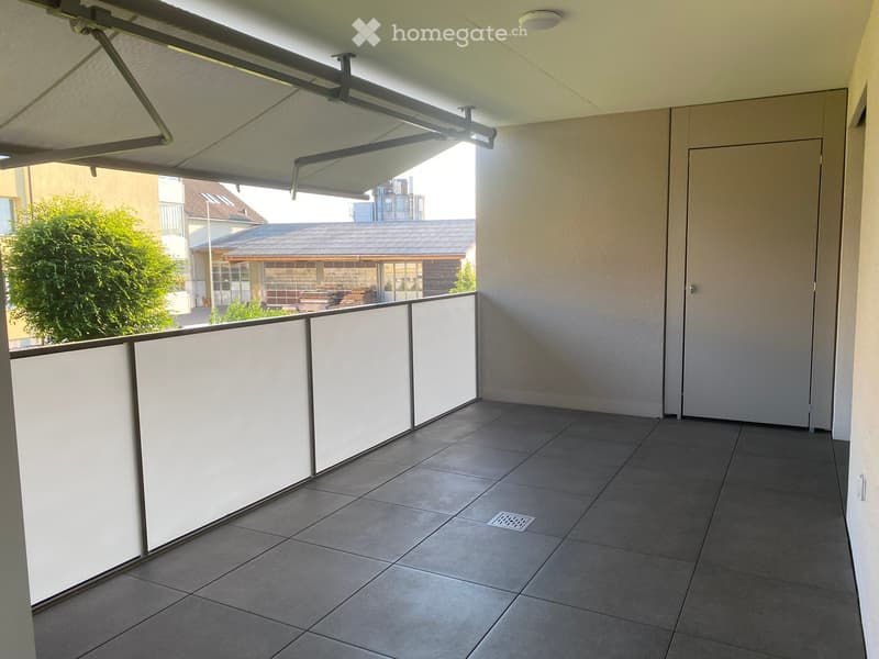 Balcony with tiled floor, white privacy screens, and a retractable awning, overlooking a residential area with trees and neighboring buildings.