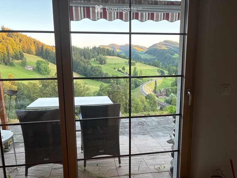 Balcony with a scenic view of green fields and hills, featuring a table and two wicker chairs.
