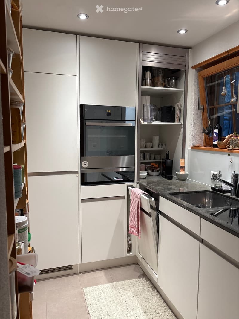 Modern kitchen with white cabinets, stainless steel appliances, and a granite countertop. Features an oven, dishwasher, sink, and various kitchen items on the counter and shelves.
