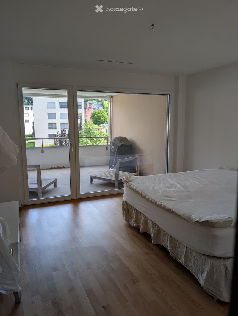Bright bedroom with wooden flooring, a neatly made bed, and a sliding glass door leading to a balcony with outdoor furniture.