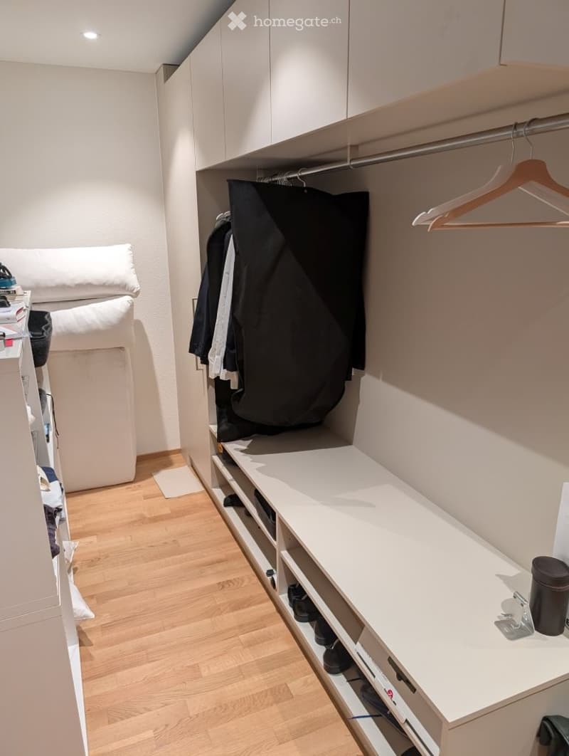 A modern, well-organized walk-in closet with white built-in wardrobes, wooden hangers, a shelf with shoes, and a small bed with pillows visible in the background.