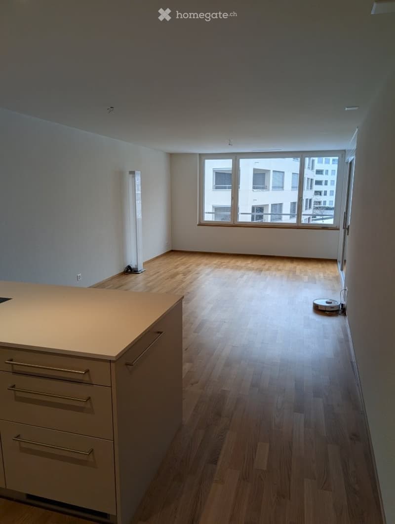 Open-plan space with a modern kitchen island and a tall floor lamp near the window. The room features wooden flooring and large windows offering a view of the city.