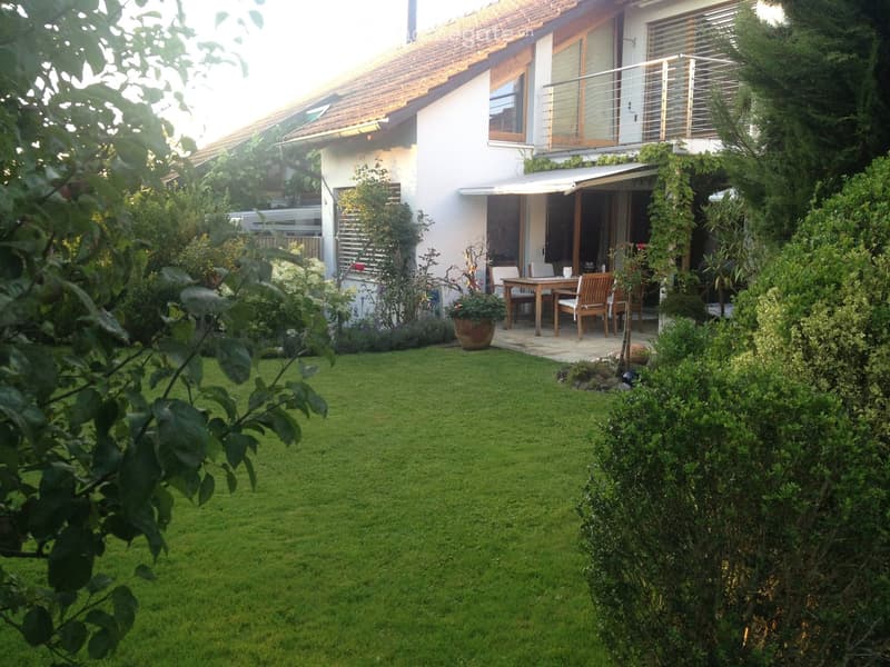 Well-maintained garden with a green lawn, wooden patio furniture, and a modern house with a balcony and large windows.