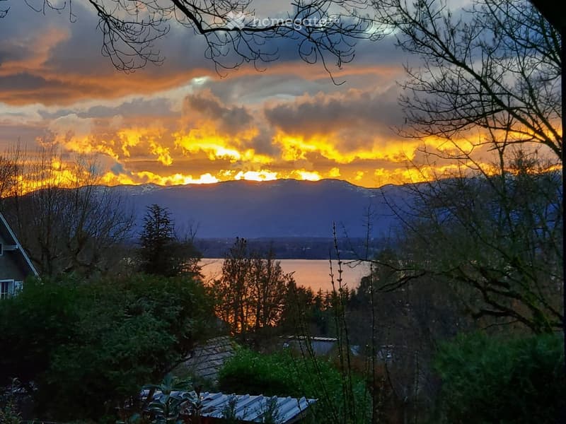 Scenic view of a lake and mountains at sunset, with trees and a house partially visible in the foreground.