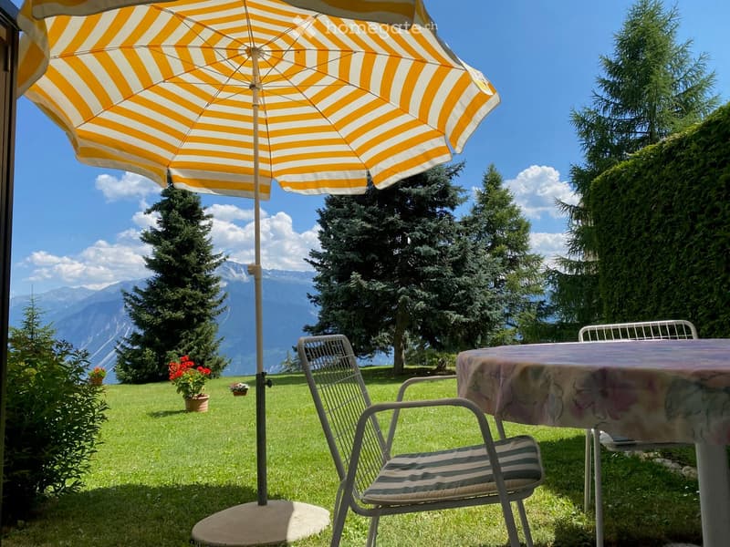 Garden with a patio umbrella, table, and chairs, surrounded by greenery and a mountain view.