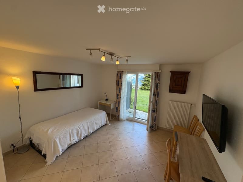 A furnished room with a bed, a small table, chairs, a mirror, a floor lamp, and a television. The room has tiled flooring and a sliding glass door leading to a grassy outdoor area.