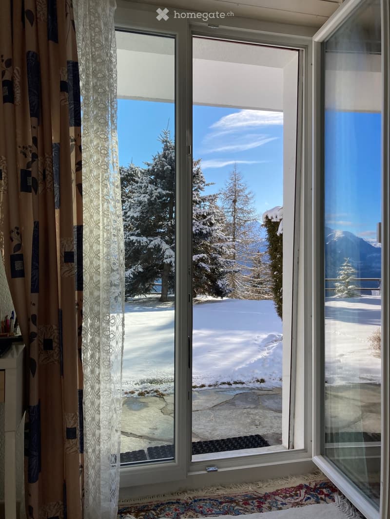 Large window with a view of snow-covered trees and mountains, partially covered by curtains and a lace panel.