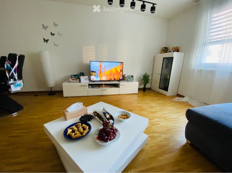 Modern living room with wooden flooring, white walls, and large windows. Features a grey sofa, white coffee table with decorative plates and bowls, a television on a white cabinet, and a floor lamp. There is also a white cabinet and a chair visible.