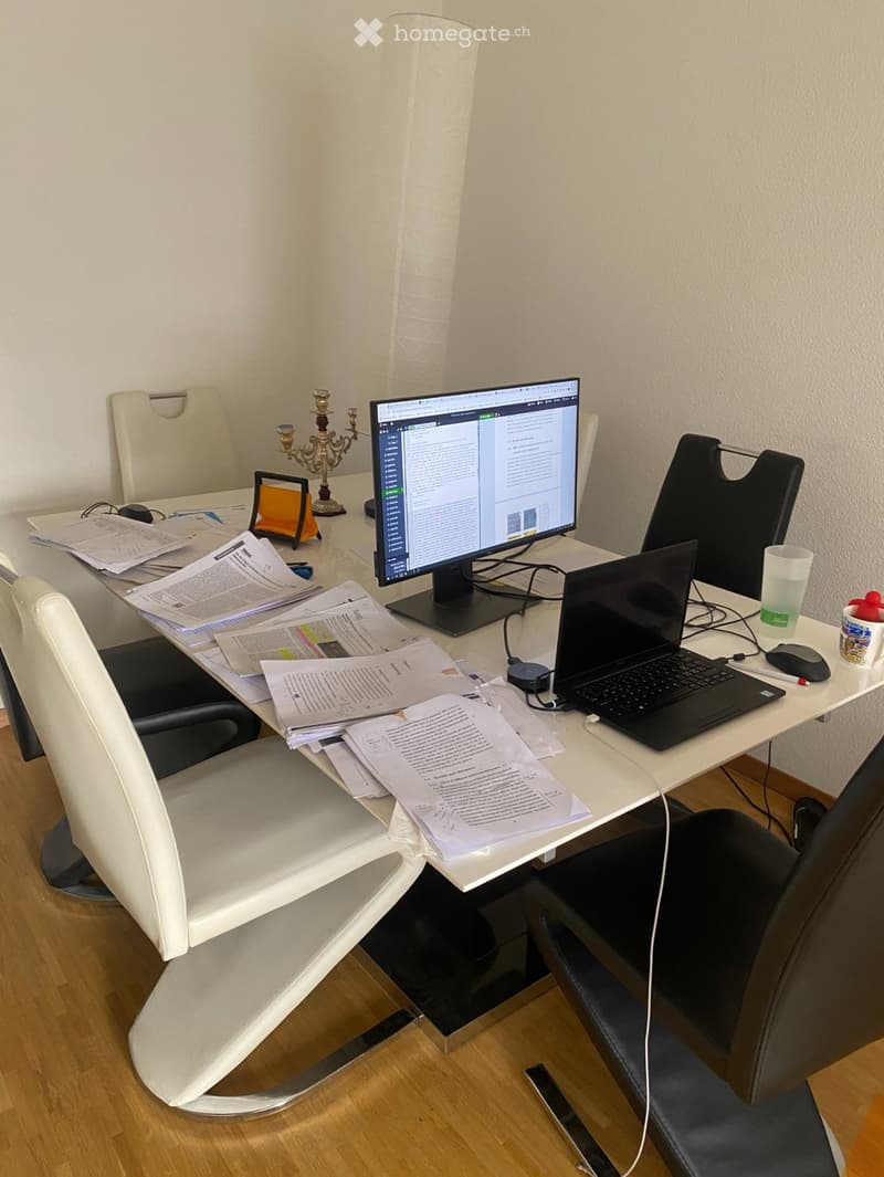A furnished room with a white table, two chairs, a laptop, a monitor, a mouse, a cup, a candle holder, and scattered papers.