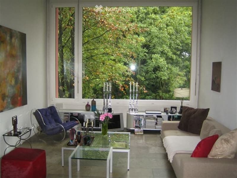 Bright living room with large windows offering a view of greenery, featuring a beige sofa with red and brown cushions, a glass coffee table, a blue armchair, a table lamp, and decorative candle holders.