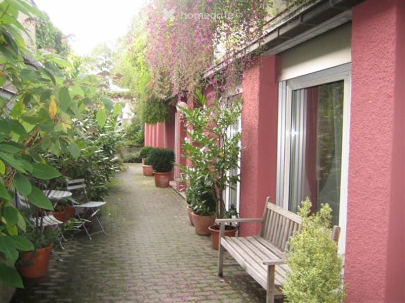 A narrow, paved outdoor corridor lined with pink walls, potted plants, and outdoor furniture including chairs and a bench.
