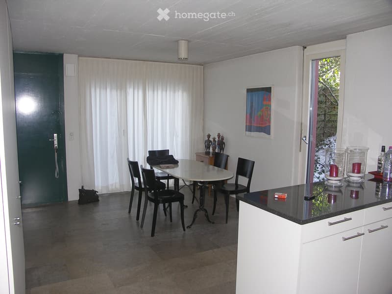 A modern, open-plan room with a dining area and a kitchen area. The dining area features a round table with four black chairs, a small side table with decorative items, and a black bag on the floor. The kitchen area has white cabinets with silver handles