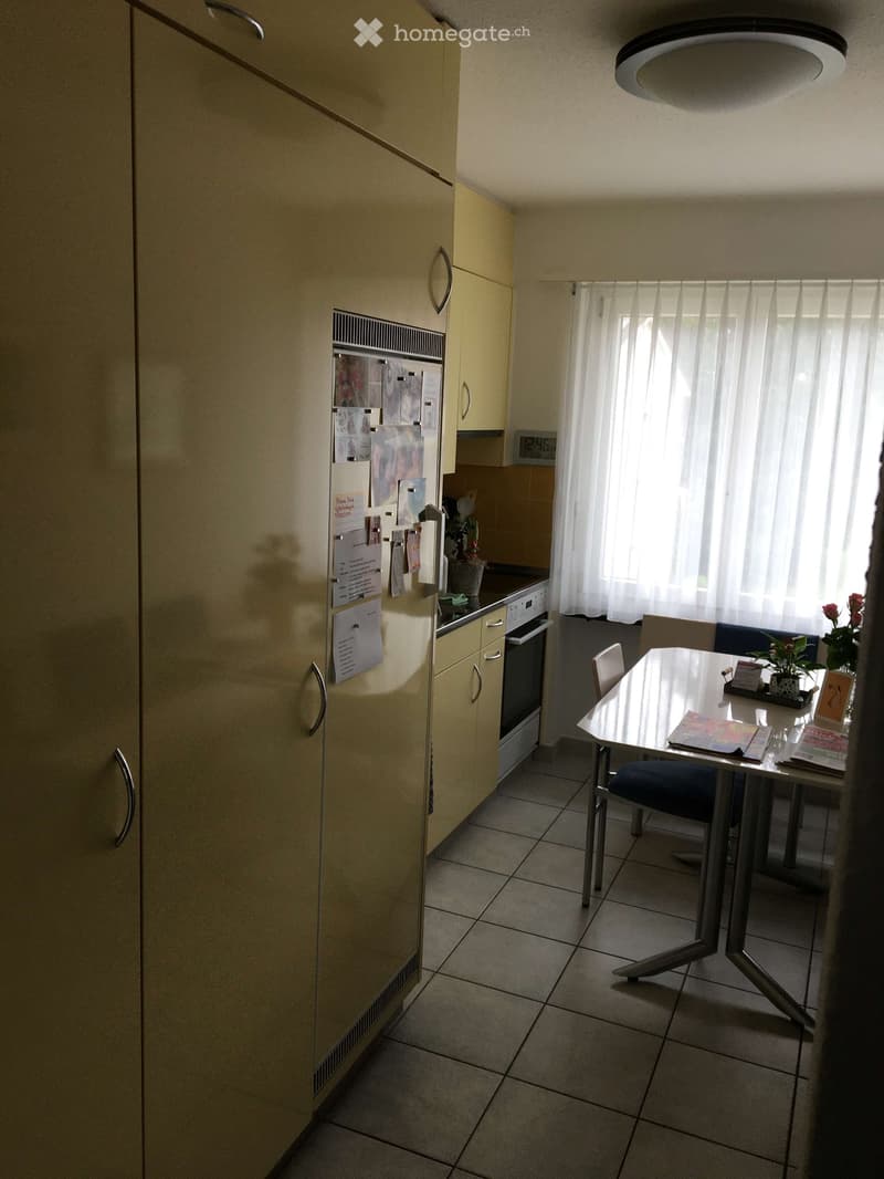 A small kitchen and dining area with light-colored cabinets, a refrigerator, an oven, a dining table with chairs, and a window with white curtains.