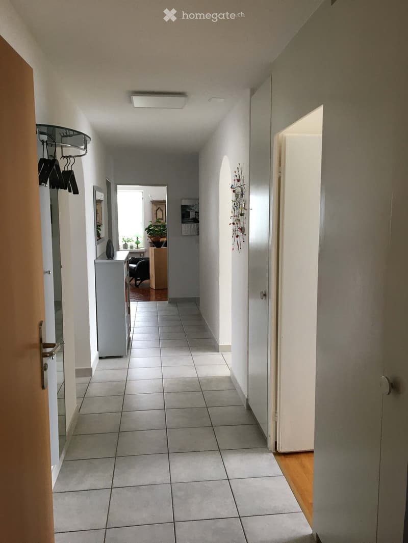 A clean, tiled hallway with white walls, a coat rack, and an open door leading to another room.