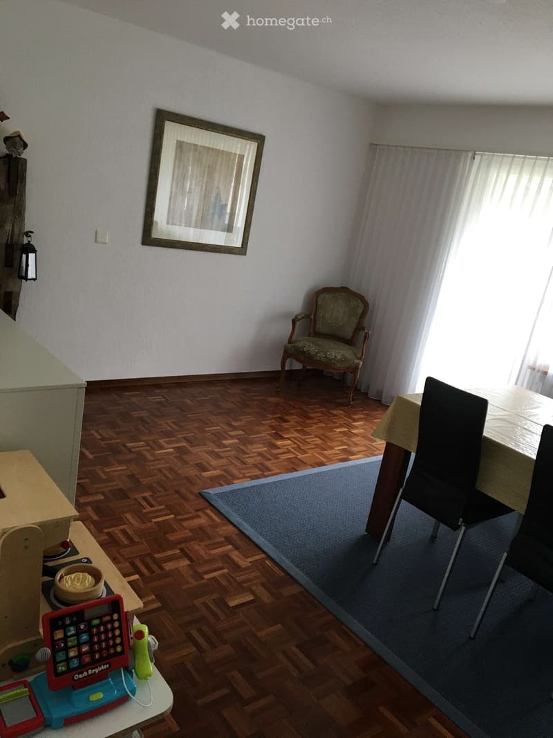 A furnished room with wooden flooring, a patterned rug, a small table with a toy cash register and bowl, a chair, and a framed picture on the wall.