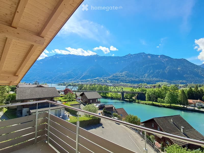 Balcony overlooking a river with a bridge, surrounded by houses and mountains under a clear blue sky.