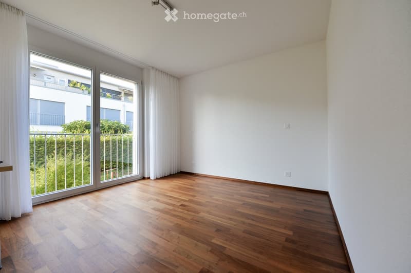 Empty room with wooden flooring, white walls, large window with white curtains, and a sliding glass door leading to a balcony.