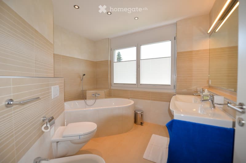 Modern bathroom with beige tiles, a bathtub, toilet, double sink, large mirror, and a window with blinds.