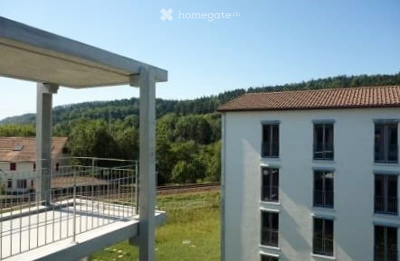 Modern multi-story building with large windows and a balcony overlooking a green landscape with trees and hills.