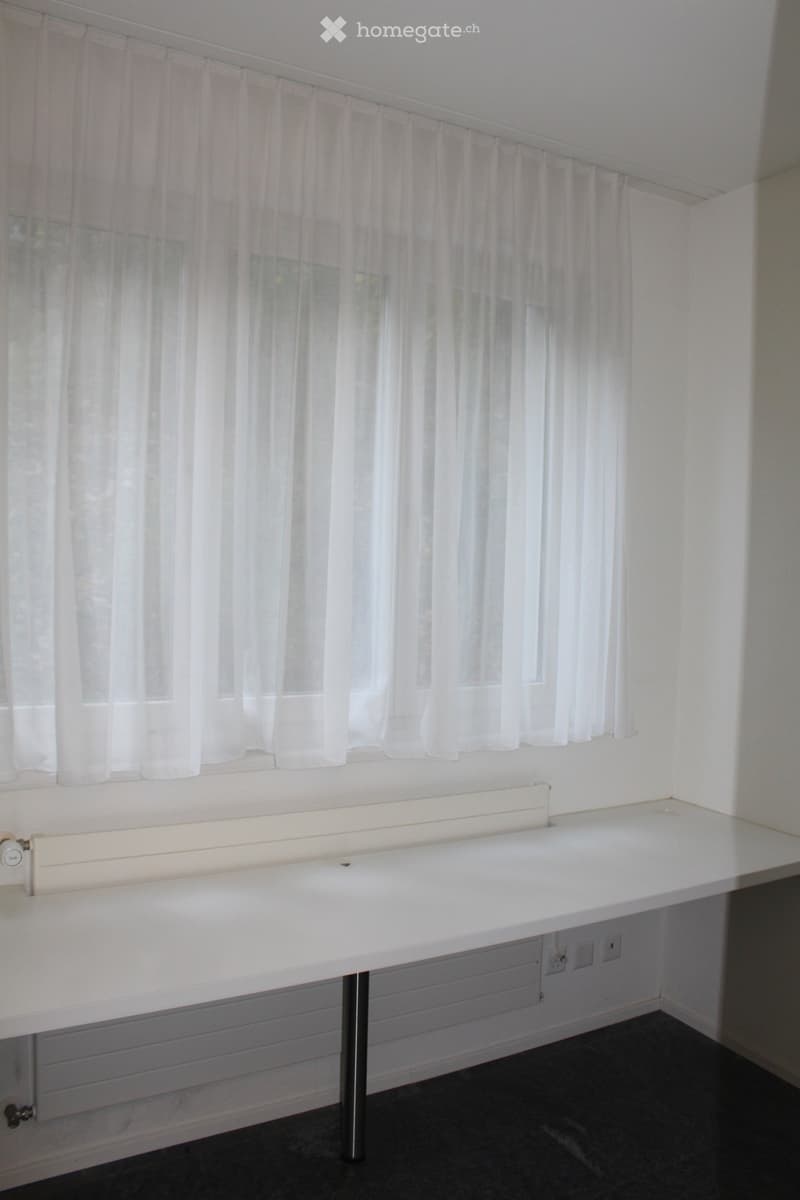 An unfurnished room with a white desk, a radiator, and white curtains on the window. The room has a dark floor and white walls.