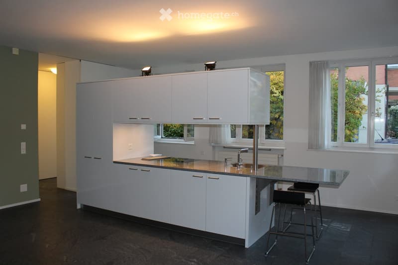 Modern kitchen with white cabinets, a central island, and a dark countertop. Two stools are placed at the island, and the space is well-lit with natural light from large windows.