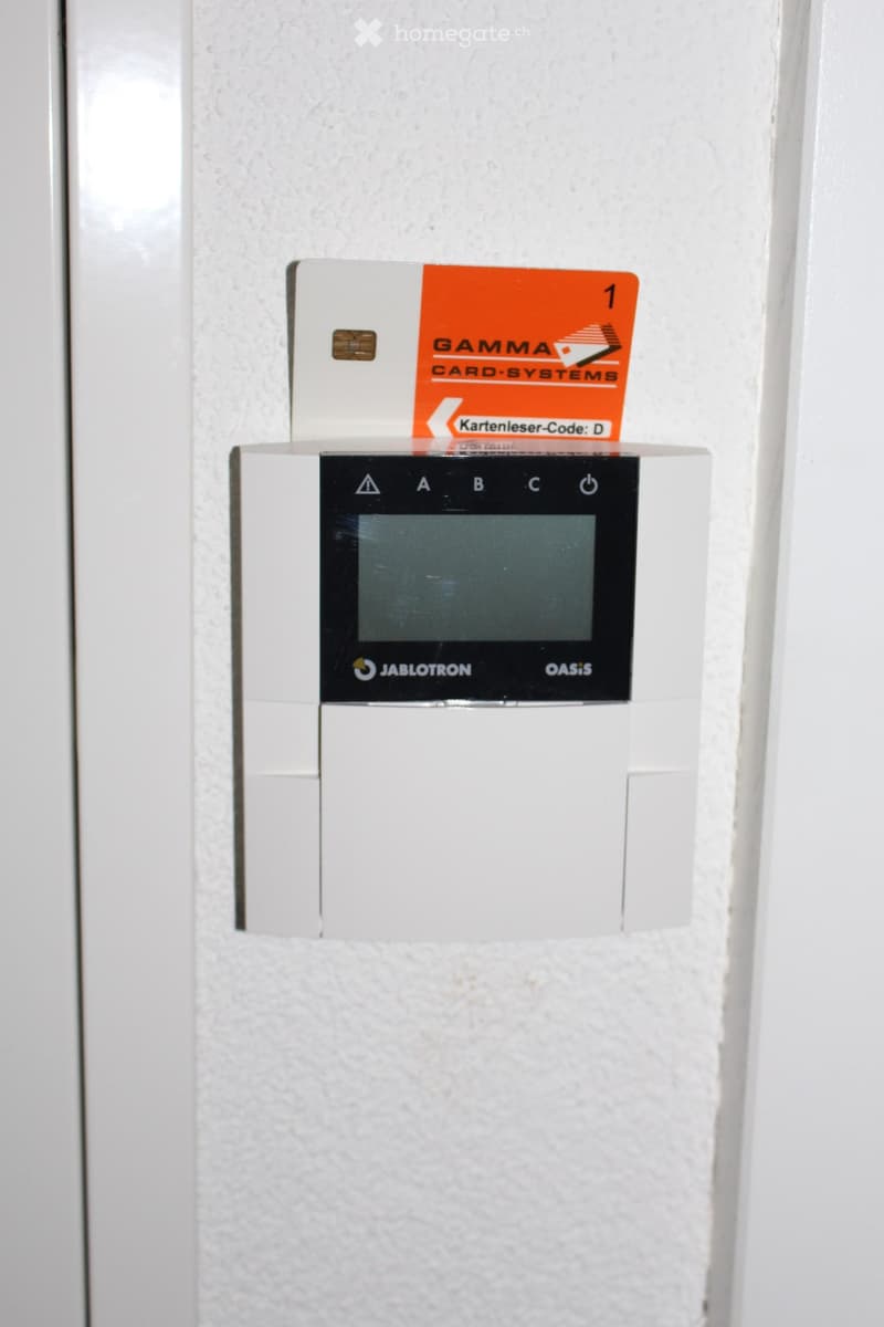A white wall with a card reader system mounted on it, featuring a small screen and buttons, with an orange card inserted above it.