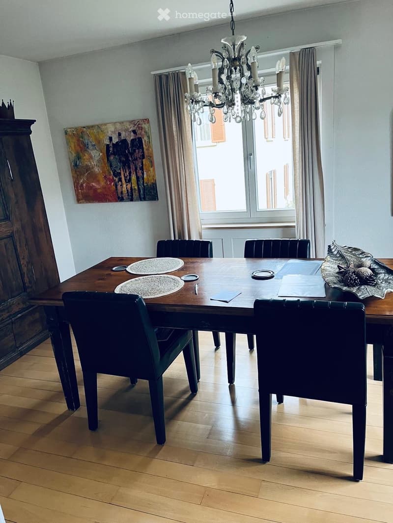 Dining room with wooden floor, chandelier, dining table with chairs, and large window with curtains.