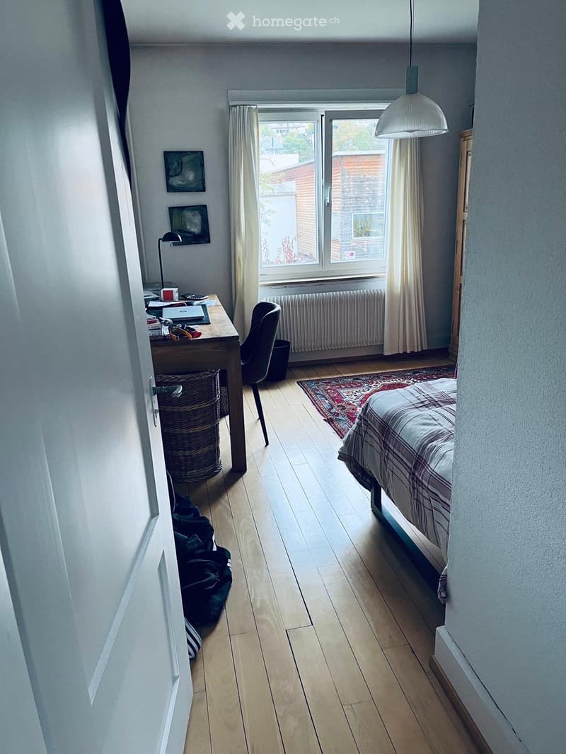 Cozy bedroom with a bed, desk, chair, laptop, wicker basket, and a large window with curtains. The room features wooden flooring and a radiator under the window.