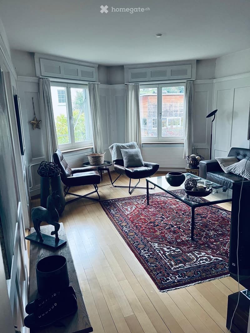 Bright living room with wooden flooring, large windows, and a patterned rug. Features a sofa, coffee table, chairs, and decorative items.